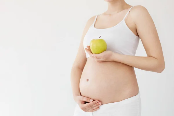 Mulher grávida segurando sua barriga e uma maçã verde saudável cheia de vitaminas esperando por seu bebê. Conceito de dieta pré-natal. Chave alta . — Fotografia de Stock