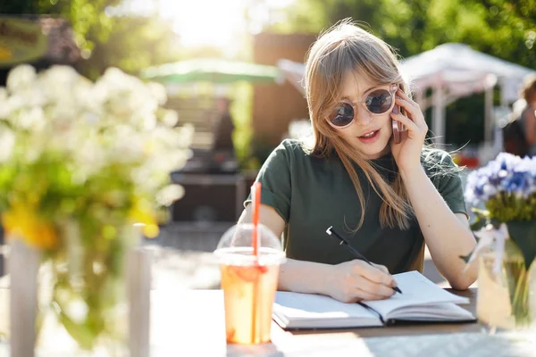 Retrato de una joven mujer de negocios o estudiante escribiendo sus planes en bloc de notas hablando en un teléfono inteligente con gafas durante una pausa en el parque en un día soleado de verano . —  Fotos de Stock