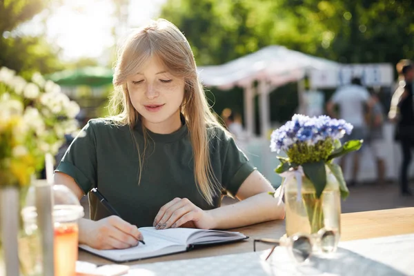 Potret seorang wanita bisnis muda atau siswa menulis rencananya di notepad yang berbicara di telepon pintar selama luch break di taman pada hari musim panas yang cerah . — Stok Foto