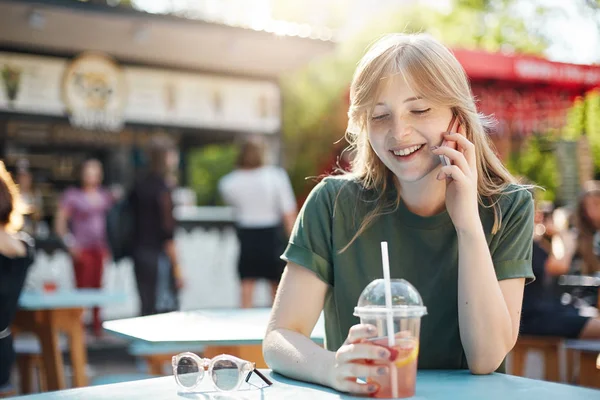 Porträt eines jungen schicken Mädchens im Gespräch mit ihrem Freund auf dem Smartphone mit Limonade in den Mittagsstunden im Park am Food Court. — Stockfoto