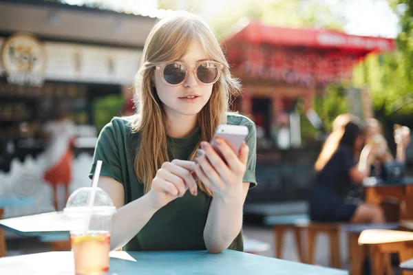 Potret wanita muda berambut pirang yang menulis sebuah pos blog di ponsel merah mudanya atau mengobrol dengan teman-teman di media sosial mengenakan kacamata pada hari yang cerah di taman . — Stok Foto