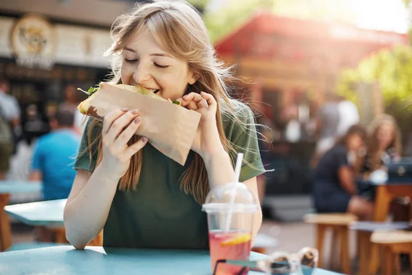 Girl eating taco. Hungry freckled blonde woman eating junk food on a food court on a sunny summer day in park.