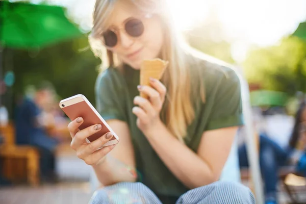 Woman eating icecream using smartphone. Portrait of happy girl with ice cream browsing through social media or messaging her friends enjoying summer in the city park wearing shades. Focus on phone. — Stock Photo, Image