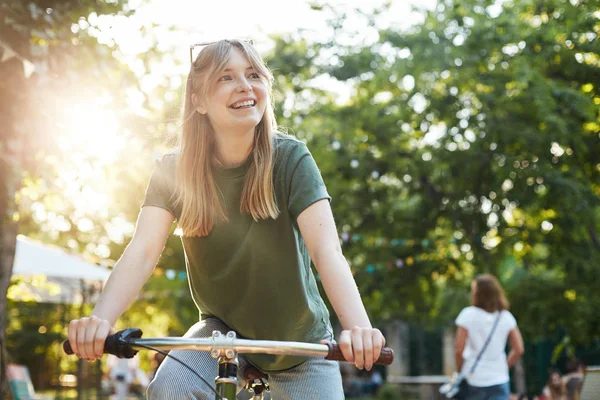 Портрет молодої красивої блондинки, насолоджуючись удаваною їздою на велосипеді в парку під час продовольчого фестивалю, посміхаючись від камери — стокове фото