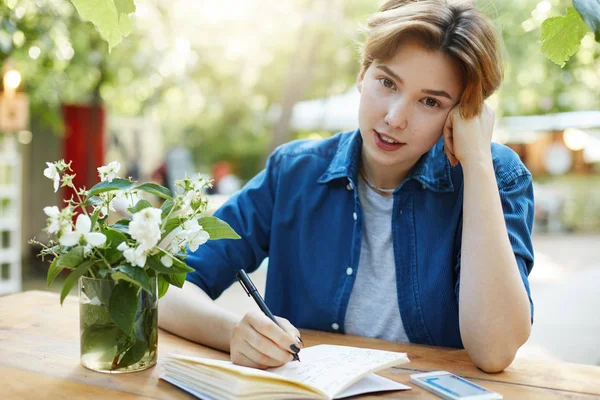 Wanita menulis di buku harian. Potret seorang gadis mengambil catatan di buku catatannya di luar ruangan merencanakan perjalanan atau liburan atau bisnis masa depan melihat kamera mengenakan kemeja biru beludru di taman . — Stok Foto