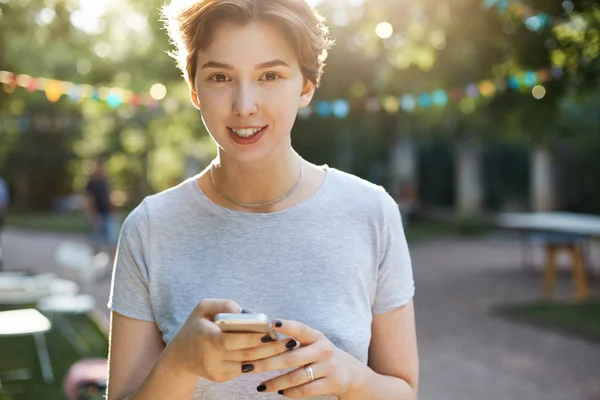 Mädchen lächelt mit Smartphone. Porträt einer jungen glücklichen Frau, die im Stadtpark lächelt und ihr Handy benutzt, in die Kamera schaut und ihrer Mutter SMS schreibt. — Stockfoto