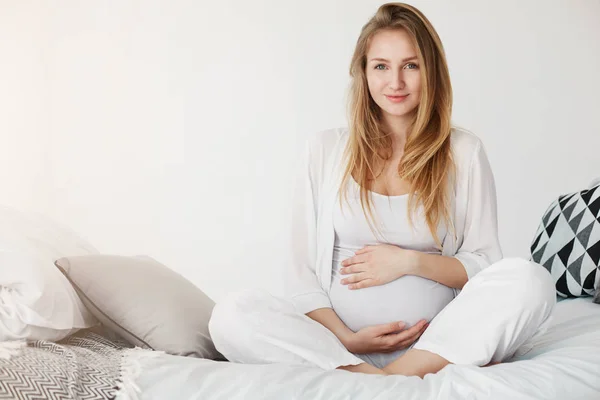 Kehamilan yang sehat. Potret seorang wanita hamil muda yang tersenyum sambil duduk di kamar tidurnya beristirahat di pagi hari sambil memegang perutnya menunggu kelahiran bayinya . Stok Gambar