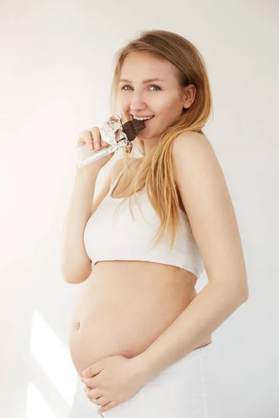 Escolha incerta entre saboroso e insalubre durante a gravidez. Mulher comendo uma barra de chocolate em seu segundo trimestre . — Fotografia de Stock
