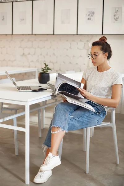 Wanita berusia dua puluhan mengejar karier dalam desain busana melihat majalah duduk di studio cerdasnya dengan komputer laptop . — Stok Foto