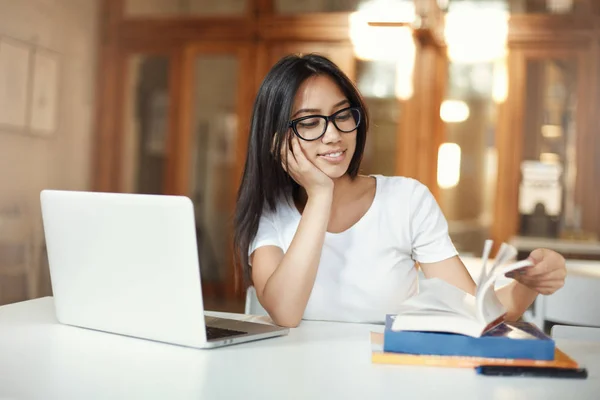 Sonriendo estudiante hembra hojeando el libro de estudio mientras trabaja en su diploma de licenciatura en una biblioteca de espacio abierto — Foto de Stock