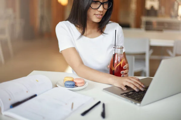 Las mujeres son inteligentes. Mujer joven usando una computadora portátil bebiendo limonada al mismo tiempo en un café, esperando para comer una pastelería macaron . — Foto de Stock