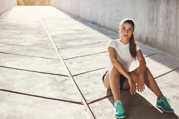 Junge Läuferin, die auf einem Bürgersteig sitzt und sich auf einen langen Lauf vorbereitet und in die Kamera blickt. Stadtsportkonzept. — Stockfoto