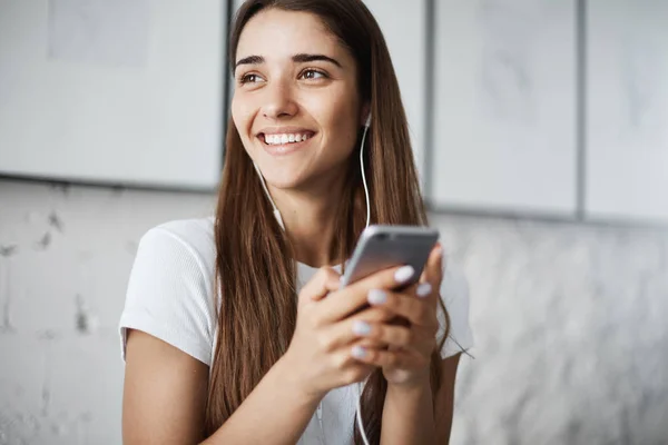 Gadis cantik berbicara di telepon menggunakan headphone kabel berpaling dari kamera tersenyum dan tertawa. Ruang kerja yang terang di latar belakang. Konsep komunikasi . — Stok Foto