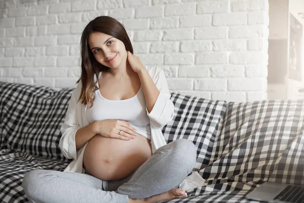 Retrato de uma jovem grávida sentada no sofá desfrutando de sua casa. Bebê sorrindo por dentro como sua mamãe faz . — Fotografia de Stock