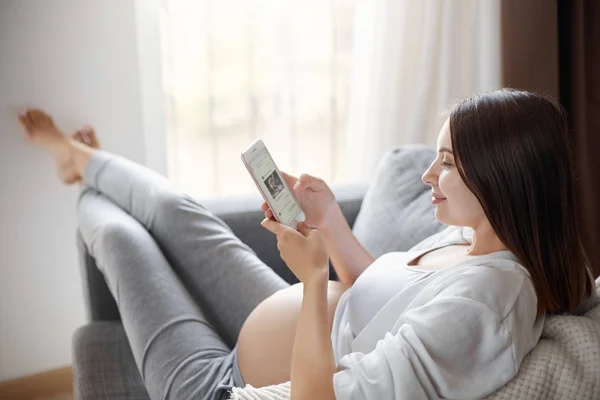 Eve onu rahat kanepe kalırken sosyal medya arkadaşları ile sohbet etmek için bir akıllı telefon kullanan genç hamile kız. Pozitif düşünme bir zorunluluktur.