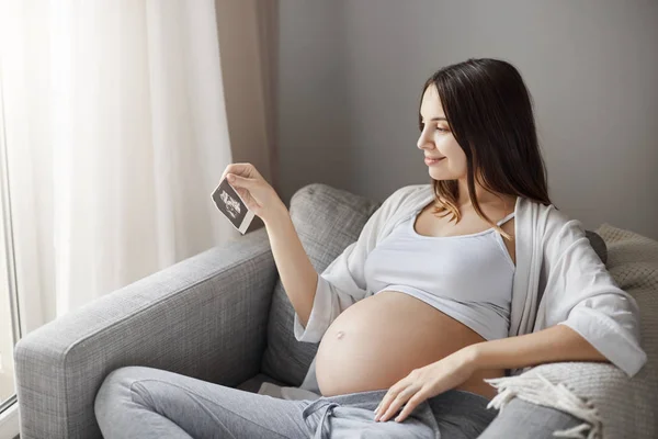 Senhora grávida olhando para uma imagem de ultra-som do bebê tentando adivinhar é um menino ou uma menina. Ficar em casa em seu sofá acolhedor . — Fotografia de Stock