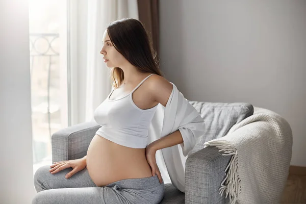 Jovem fêmea grávida esfregando as costas para aliviar as dores. Ficar em sua casa acolhedora e acolhedora passar as últimas semanas de gravidez . — Fotografia de Stock