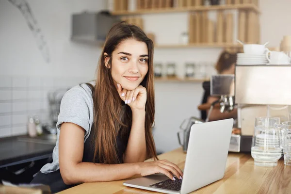 Propietaria de café femenino usando computadora portátil esperando al primer cliente por la mañana mirando a la cámara sonriendo . — Foto de Stock