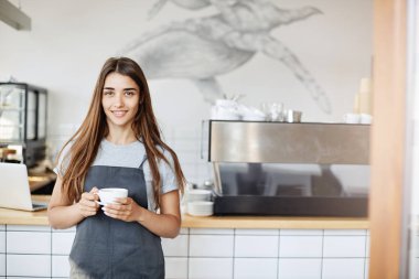 Kahve makinesi cappucino bardak tutan kameraya bakarak yakın kafe ve hamur işi Dükkanı ayakta çalışan genç bir kadın portresi.