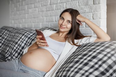 Genç anne gebelik hakkında daha fazla keşfetmek için bir akıllı telefon kullanarak. Bebeğini evde bekleyen rahatlatıcı. Sağlıklı yaşam konsepti.
