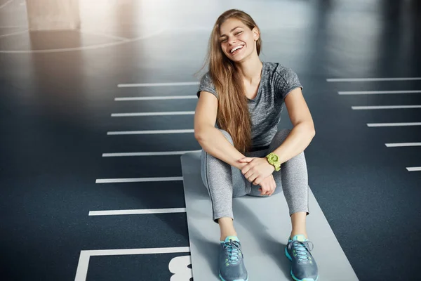 Portrait of laughing young female fitness trainer sitting in empty space after a long hard workout. Fitness concept. — Stock Photo, Image