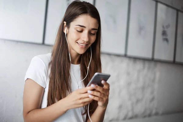 Chica feliz usando un teléfono inteligente escuchando mensajes de voz o viendo videos de gatos en auriculares blancos. Trabajador perezoso . —  Fotos de Stock