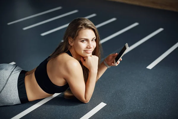 Retrato de mulher bonita deitada no ginásio depois de um treinamento duro usando um telefone inteligente para postar fotos nas mídias sociais . — Fotografia de Stock