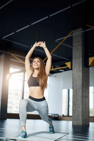 Beautiful female fitness idol making squats smiling early in the morning in empty gym. Portrait of fit woman training hard. — Stock Photo, Image