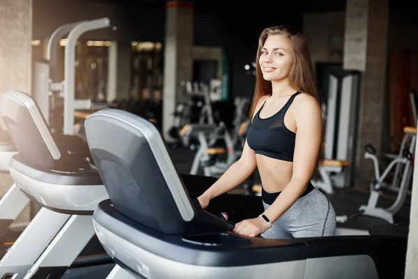 Portrait of crazy fit woman on a threadmill smiling looking at camera. Athletic coach warming up early in the morning in empty gym. — Stock Photo, Image