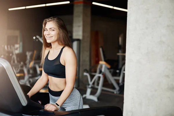 Junge weibliche Körper-Fitness-Trainerin, die auf einem Laufband läuft und lächelnd in die Kamera blickt, die mit einem Monitor die Herzfrequenz verfolgt. Athletiktrainer wärmt sich früh in leerer Turnhalle auf. — Stockfoto