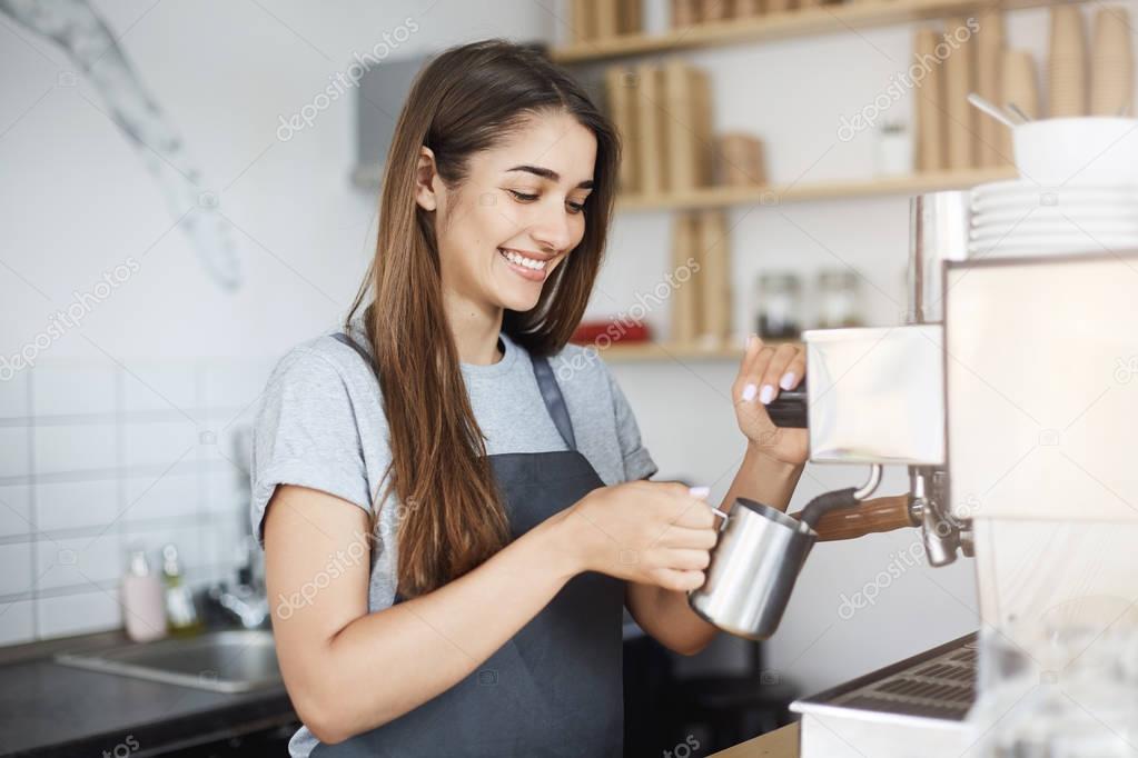 Young woman steaming milk to make morning capuccino coffee in bright cafe laughing and smiling cheerfuly.