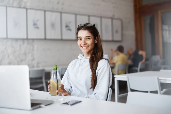 Linda atractiva mujer joven europea sentada frente a la computadora portátil, con sudadera de moda y gafas elegantes, beber bebidas mientras está en la cafetería. Freelancer se tomó un descanso del trabajo, bebiendo cóctel — Foto de Stock