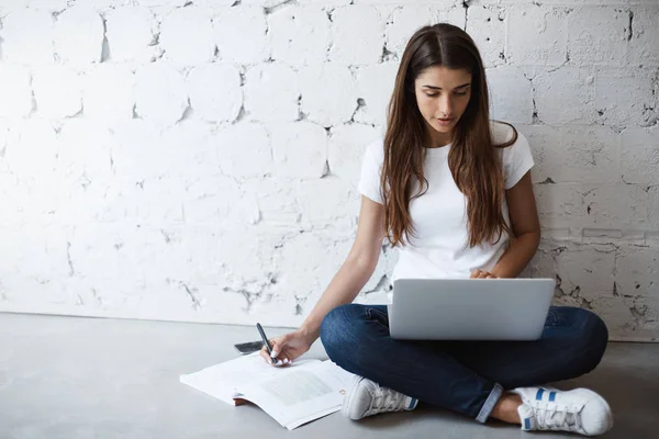 Portretu inteligentnego zajęty kobiety, robiąc notatki, podczas gdy pochylony na ścianie i siedzi na podłodze z laptopa na skrzyżowane nogi. Dziewczyna ma zwyczaj do pracy lub nauki wszędzie, ale nie przy stole. — Zdjęcie stockowe