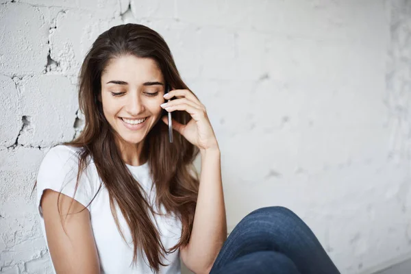 Portrait d'une heureuse femme européenne souriante assise sur le sol et appuyée sur un mur de briques blanches tout en parlant sur un smartphone avec un bon ami. Dieu merci, nous avons wifi presque partout . — Photo