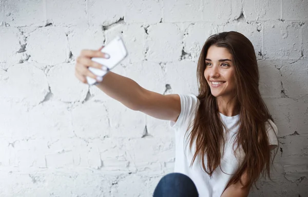 Портрет очаровательной европейской модели, делающей селфи на смартфоне возле белой кирпичной стены, весело улыбающейся. Модный блогер делает фото, чтобы разместить его в своем блоге. У неё много поклонников. — стоковое фото