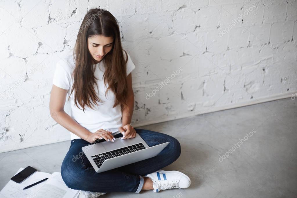 Upper view of creative young female, using laptop while sitting on floor, leaning on wall, surrounded with gadgets and books. Designer works on new project she needs to present tomorrow.