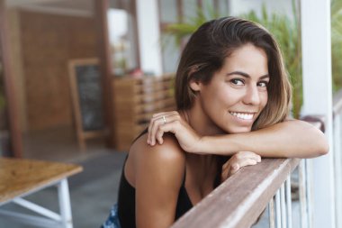 Wellbeing, beauty and tourism concept. Happy attractive young hispanic woman lean on terrace handrail, smiling and looking camera dreamy, contemplate outside nature, travel to relax clipart