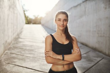 Rekorunu kırabilir misin? Siyah spor sutyen ve şort giymiş motive ve kendinden emin bir sporcu, spor takip cihazı takıyor, kolları çapraz güvenli kamera takıyor yaz güneşi altında sabah koşuyor.