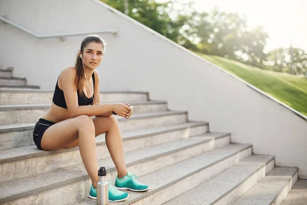 Corredor femenino activo y saludable comenzar el día bien, tomar aliento después de la sesión de entrenamiento productivo al aire libre, sentarse en la escalera con botella de agua, escuchar música, terminar jogging mañana mirada satisfecho — Foto de Stock