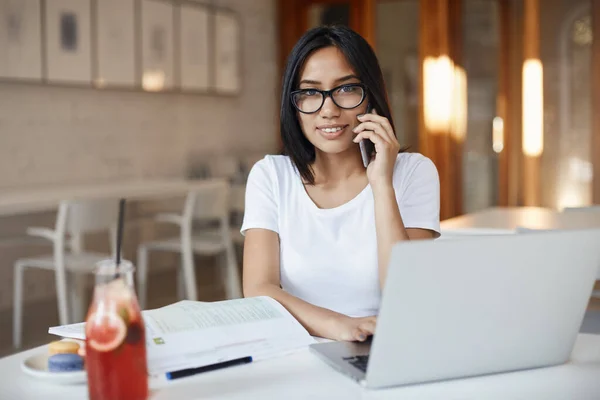 Pensive and smart female entrepreneur working remote from cafe, using laptop and calling customer via smartphone, taking notes with pen, drink orange juice, feeling productive and energized Royalty Free Stock Photos