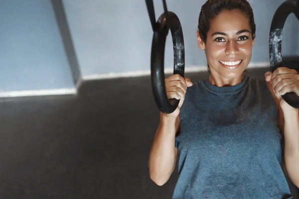 Upper-angle shot smiling sportswoman, female athlete in sportswear pumping biceps, push-up using gym equipment, hanging on rings and grinning, look camera, relaxed with sweat on forehead — Stock Photo, Image