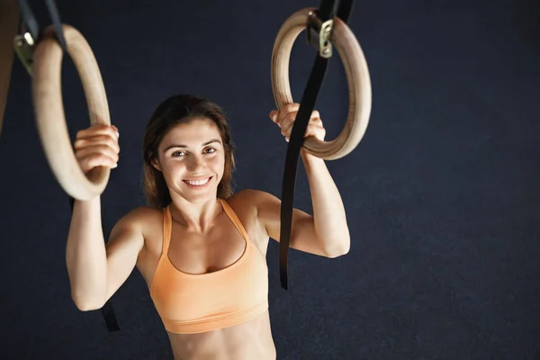 Upper-angle shot smiling motivated attractive young woman in orange sportsbra, grip gymnastics ring, grinning camera, pull herself up, pumping muscles, training in fitness club — Stock Photo, Image