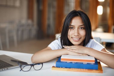 Refah, genç yaşam tarzı, online eğitim konsepti. Gülümseyen güzel bir kadın notların ve kitapların üzerine uzanmış kamerayı neşeyle izliyor, çalışma alanından uzakta çalışıyor, laptopla kafede ders çalışıyor.