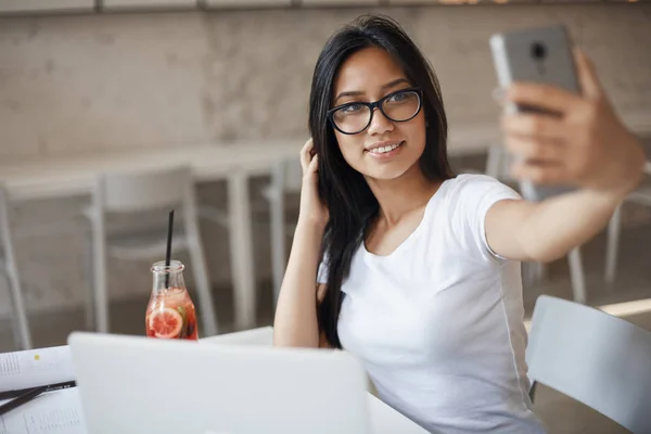 Schöne junge Studentin mit Brille, die während des Studiums ein Selfie mit dem Smartphone macht, Hipstermädchen, die Online-Beiträge über E-Learning macht, Saft trinkt und Notizen für einen Aufsatz an der Universität macht — Stockfoto