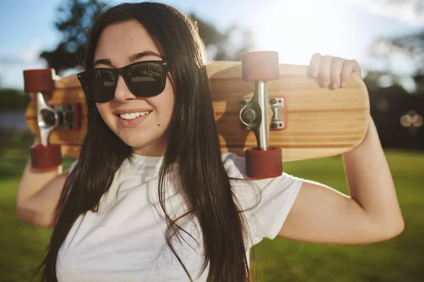 Friluftsliv, kommunalt skateopplegg. Nær-opp attraktiv skater jente i solbriller, hvit t-skjorte står i grønn park, sol skinner bak, kjøler i solstråler, holder tre penny board – stockfoto