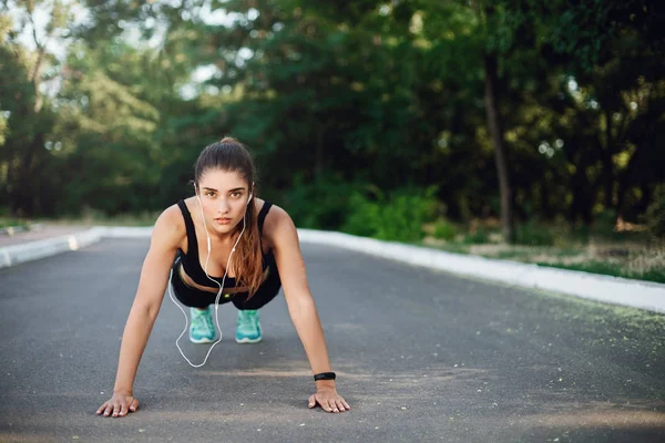 Intrygująca młoda, zdeterminowana kobieta sportowa w stroju aktywnym stojąca na betonowej drodze w parku, wykonująca ćwiczenia push-up podczas porannego rozgrzewki przed joggingiem, słuchająca muzyki przewodowe słuchawki — Zdjęcie stockowe