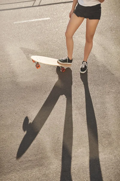 Vertikalt skott kvinna ben i shorts och sneakers lyfta upp skateboard, skridskoåkare flicka ridning penny board njuta av sommaren kväll aktiv aktivitet, öva nya tricks longboard, utföra stunts — Stockfoto