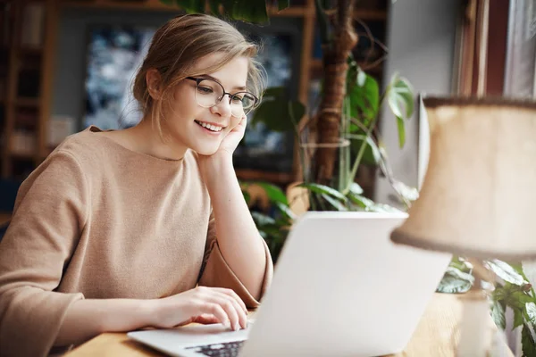 Alegre chica caucásica joven en gafas usando el ordenador portátil, compras en línea o navegar por Internet, charlando con amigos, editar fotos para el proyecto universitario, sonriendo como pantalla de ordenador que mira — Foto de Stock