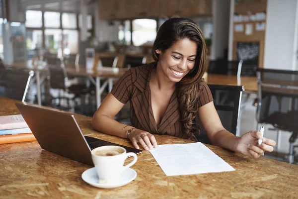 Affari, concetto di economia gig, Affascinante giovane imprenditrice brasiliana seduta co-working caffè, lettura di documenti di lavoro, prendere appunti, utilizzando il computer portatile come lavorare a distanza dall'ufficio, bere cappuccino — Foto Stock