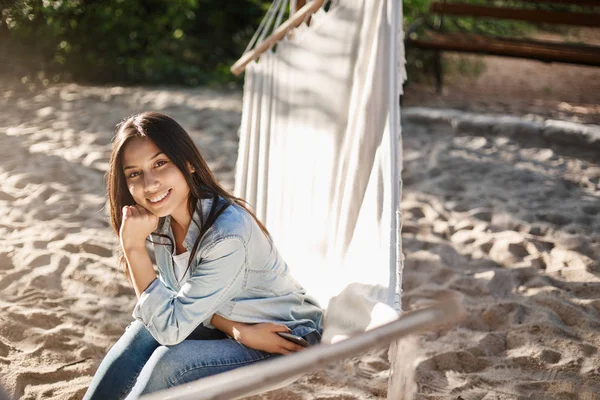 Día de verano perfecto, ocio de fin de semana y concepto de diversión. Hermosa chica sonriente sentada en la hamaca empapando los pies en la arena de la playa, sonriendo con alegría la cámara, relajarse en el parque local, disfrutar del día libre —  Fotos de Stock